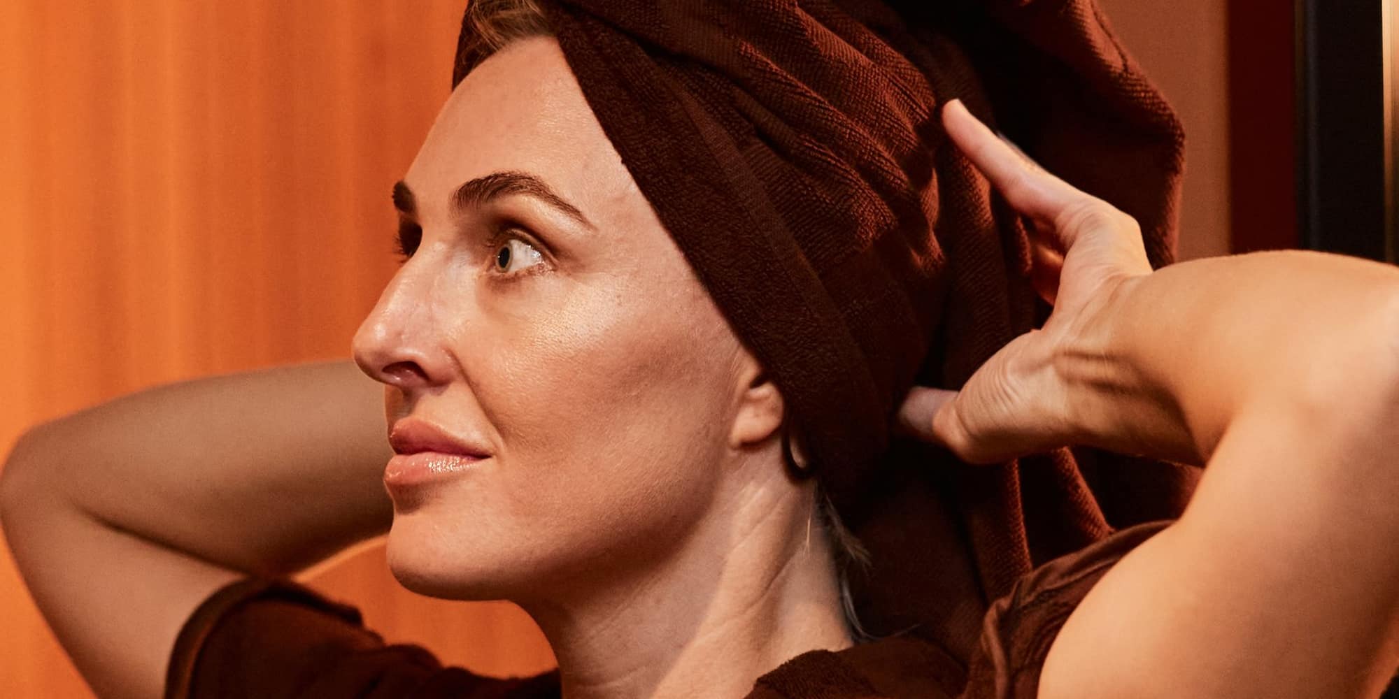 A woman in a dark robe with her hair wrapped in a towel turban, lit by warm sauna light.