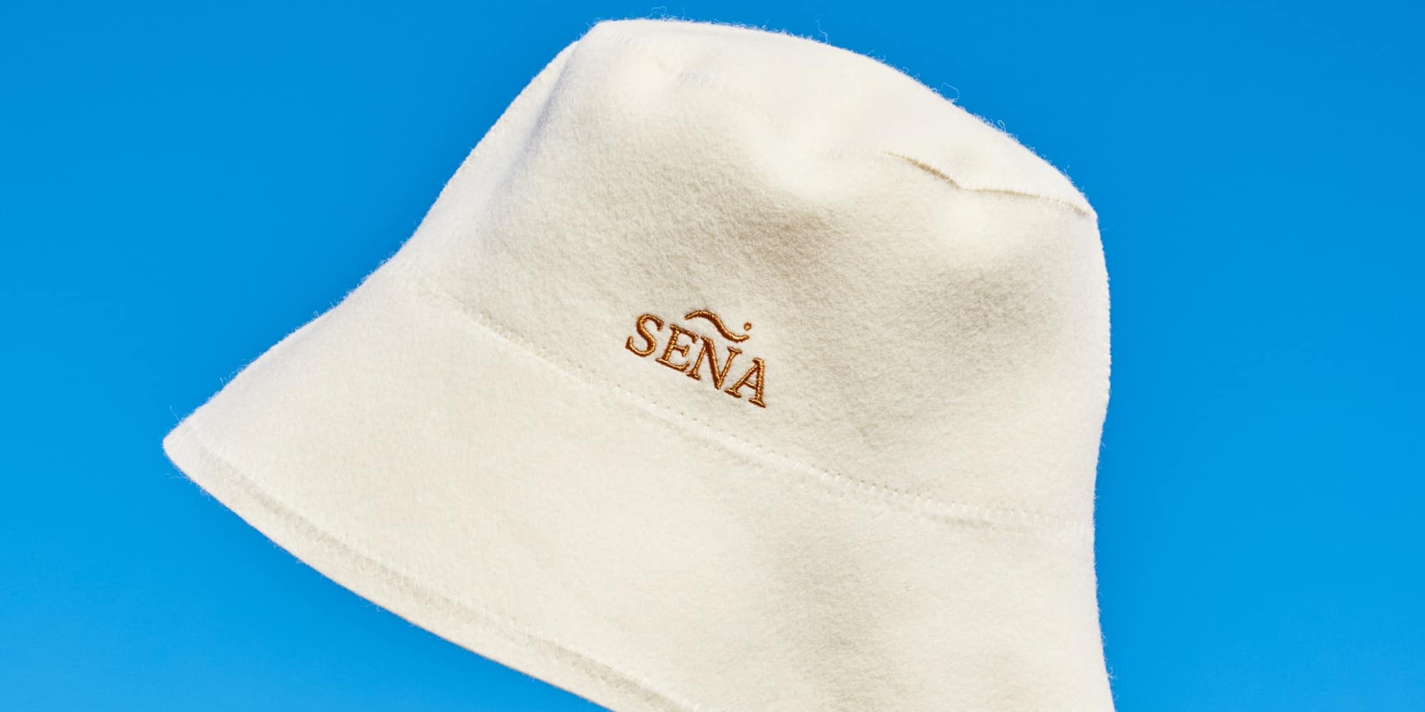 A cream merino wool SENA sauna hat photographed against a clear blue sky.