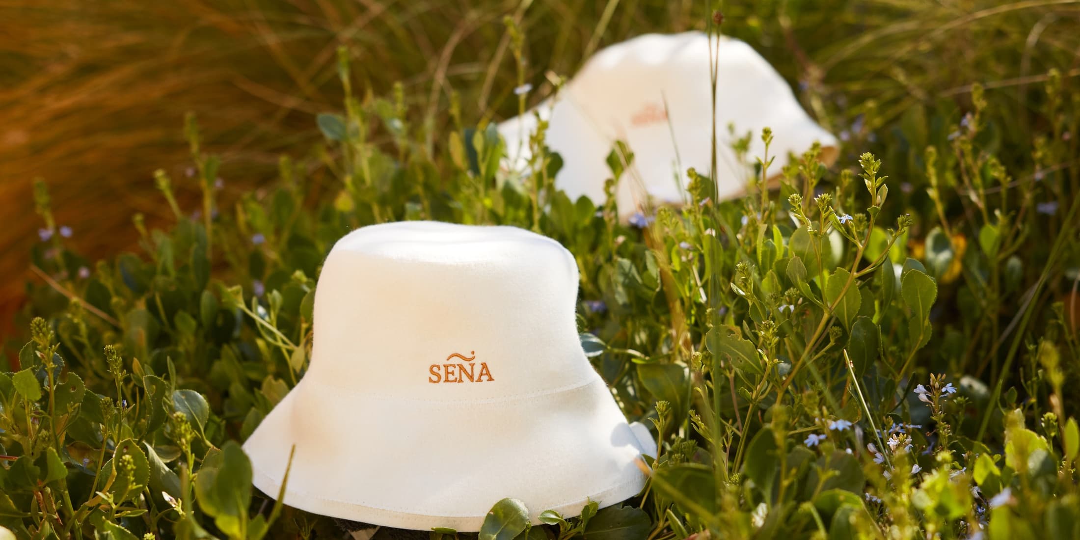 A cream merino wool SENA sauna hat resting in coastal scrub in bright daylight, the felted wool surface clearly visible.